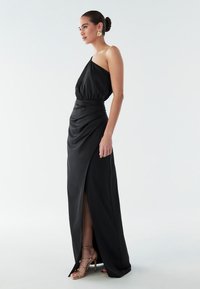 Schwarzes Abendkleid mit einem Schulterträger, gerafftem Oberteil und drapiertem Rock. Es hat einen hohen Schlitz und einen glatten Stoff. Goldene Riemchen-Heels runden den Look ab.
