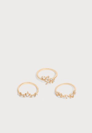 Trois bagues en or avec de petits accents en cristal clair disposées sur un fond gris clair.