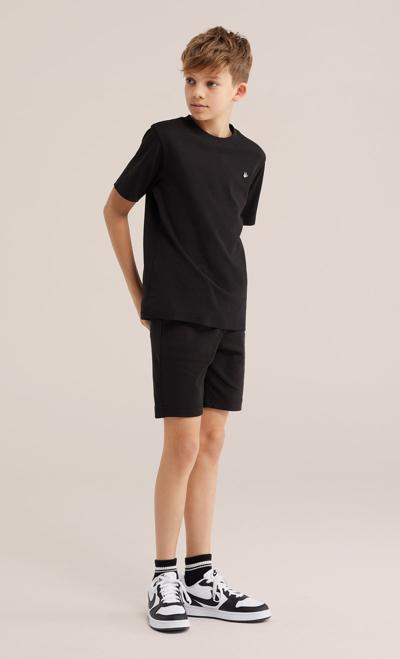 Garçon debout, les mains derrière le dos, portant un t-shirt noir, un short noir, des baskets noires et blanches, regardant sur le côté sur un fond uni.