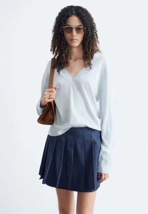 Femme aux cheveux bouclés portant des lunettes de soleil, un pull bleu clair à col en V, une jupe plissée bleu marine et un sac à bandoulière marron.