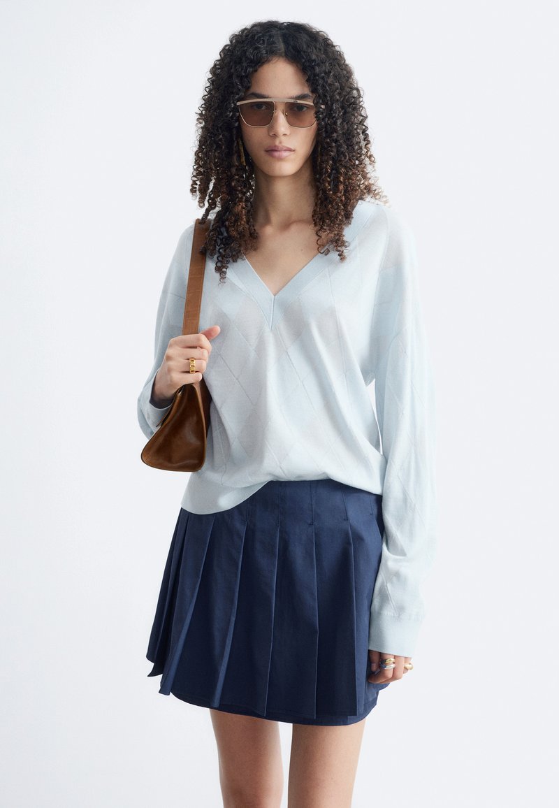 Femme aux cheveux bouclés portant des lunettes de soleil, un pull bleu clair à col en V, une jupe plissée bleu marine et un sac à bandoulière marron.