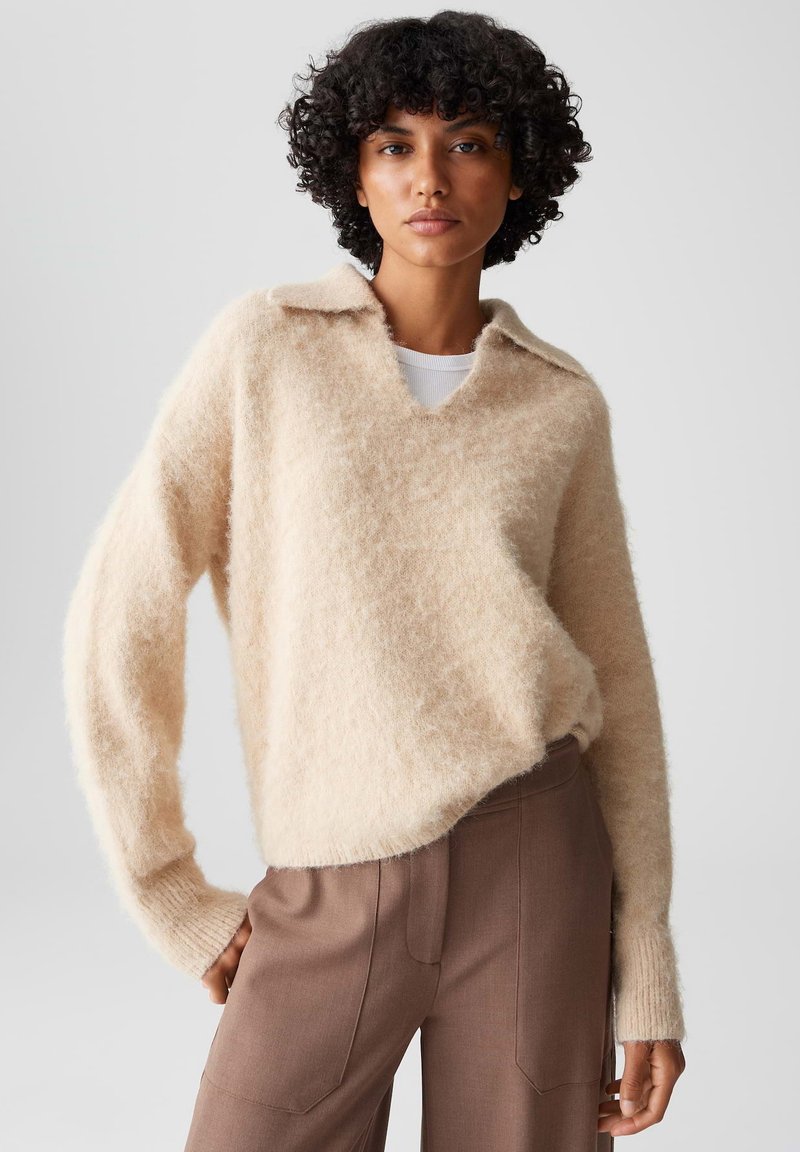 Maglione di lana beige, morbido e fluffy, con scollatura a V e colletto. Presenta una vestibilità ampia e maniche lunghe, abbinato a pantaloni marroni a vita alta.