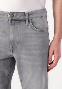 Pilkos džinsinio audinio kelnės su penkių kišenėlių dizainu, metaline sagtimi ir subtilia tekstūra. Turi vidutinio aukščio juosmenį ir tiesios kojos siluetą.