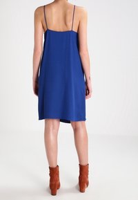 Vestito blu senza maniche con spalline sottili, design fluido, lunghezza a metà coscia, abbinato a stivaletti con tacco marroni alla caviglia. Tessuto dalla texture liscia.