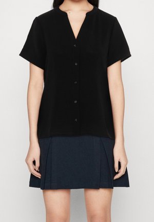 Femme portant un chemisier noir à manches courtes boutonné et une jupe plissée bleu marine foncé, debout devant un fond uni.