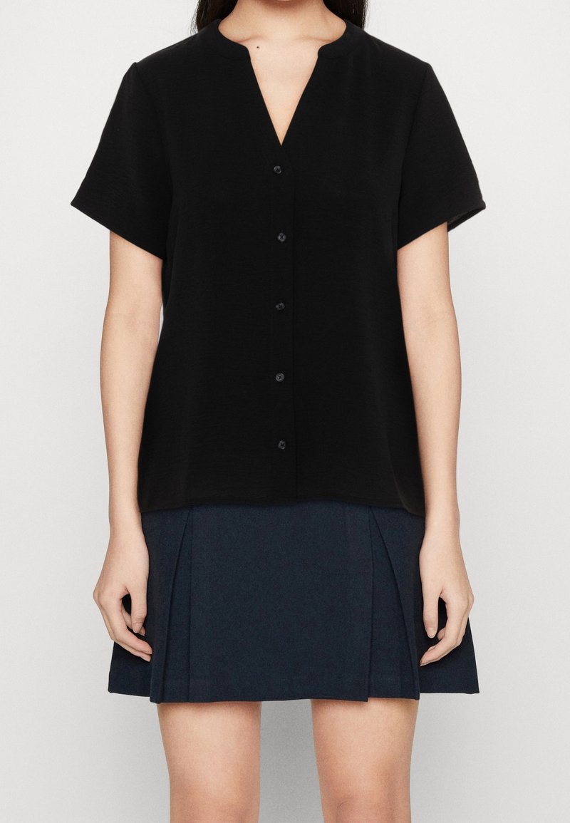 Femme portant un chemisier noir à manches courtes boutonné et une jupe plissée bleu marine foncé, debout devant un fond uni.