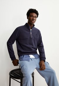Sweatshirt zippé bleu marine avec un col montant et une couture sur la poitrine, associé à un pantalon cargo bleu clair. Détail de logo minimal sur la poitrine.