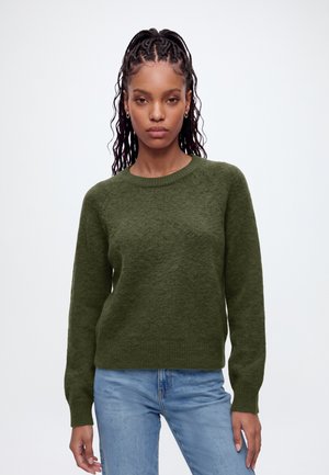 Femme aux cheveux tressés portant un pull en maille vert et un jean bleu, debout devant un fond clair uni.