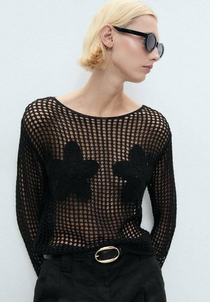 Schwarzer Strickpullover mit Netzdesign, ausgestattet mit übergroßen schwarzen Blumenapplikationen auf der Vorderseite. Das Model trägt Sonnenbrillen und hochtaillierte Hosen.