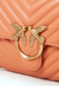 Korallfärgad läderhandväska med chevron-quiltning, med en guldfärgad spänne i form av två fåglar. Märkesnamnet "PINKO" syns på metallbeslagen.