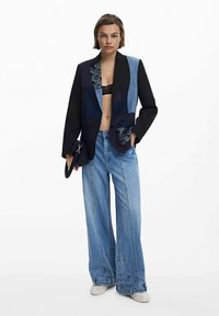 Asymmetrischer Blazer aus schwarzen und blauen Stoffen, mit floralen Akzenten und einer Tasche. Kombiniert mit weit geschnittenen Jeans in Hellblau.