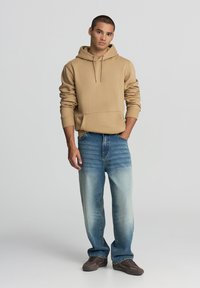 Felpa beige con tasca a marsupio e cordino regolabile, abbinata a jeans in denim chiaro con finitura sbiadita e vestibilità ampia.