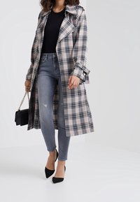 Abrigo de trench a cuadros en beige, negro y rosa; diseño abotonado con puños con cinturón. Combinado con jeans skinny grises y tacones puntiagudos negros.