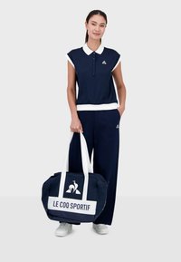 Conjunto deportivo azul marino con acentos blancos, que incluye una camiseta con cuello y pantalones holgados. Una gran bolsa azul marino muestra "LE COQ SPORTIF" en blanco.
