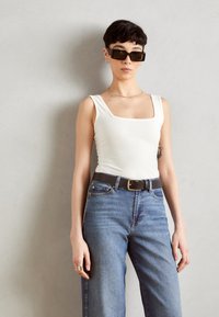 Top sans manches blanc ajusté avec un décolleté carré, associé à un jean taille haute bleu clair. Ceinture noire avec boucle en métal, lunettes de soleil et cheveux courts.