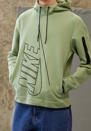 Osoba ubrana w jasnorozową bluzę z kapturem Nike z czarnymi troczkami i dużym czarnym logo Nike na konturze, w połączeniu z niebieskimi dżinsami. Ręce w kieszeniach.