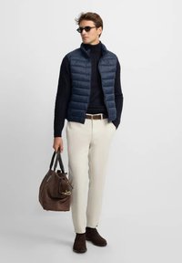 Hombre con gafas de sol, chaleco azul marino y suéter, pantalones crema, zapatos marrones, sosteniendo un bolso de cuero marrón, de pie contra un fondo blanco.