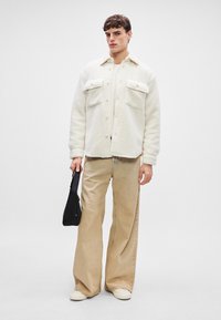 Hvit fleecejakke med to brystlommer, kombinert med lysebeige vide bukser og hvite sko. Bærer en liten svart veske.