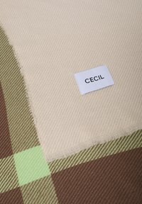 Beige tæppe med diagonalt stribet mønster, der har grønne og brune ternede accenter samt en hvid etiket med teksten "CECI L". Flosset kanter.