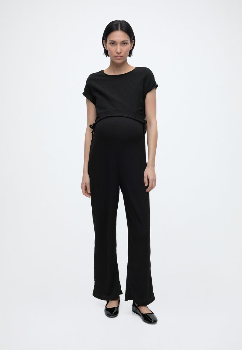 Έγκυος γυναίκα με κοντά μαύρα μαλλιά, φοράει μαύρο κοντομάνικο μπλουζάκι, φαρδιά μαύρα παντελόνια και μαύρα παντόφλες σε λευκό φόντο.