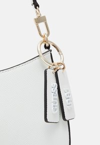 Bolso blanco texturizado con detalles en negro, herrajes dorados y etiquetas con el logo que muestran "GUESS" y "1981". Presenta un diseño liso y elegante.