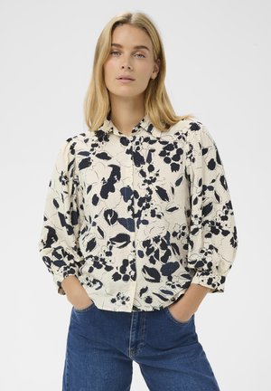 Femme blonde les mains dans les poches portant un chemisier crème à imprimé floral noir et un jean bleu, devant un fond uni.