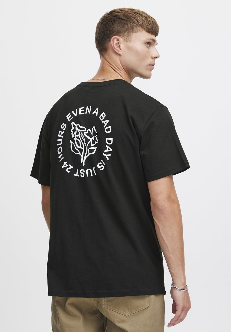 Sort bomulds t-shirt med hvid blomster grafik og teksten "SELV EN DÅRLIG DAG ER BARE 24 TIMER" på ryggen. Korte ærmer, afslappet pasform.