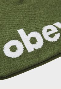 Olive green knitted beanie with large white embroidered text. Text features rounded shapes and a soft texture.