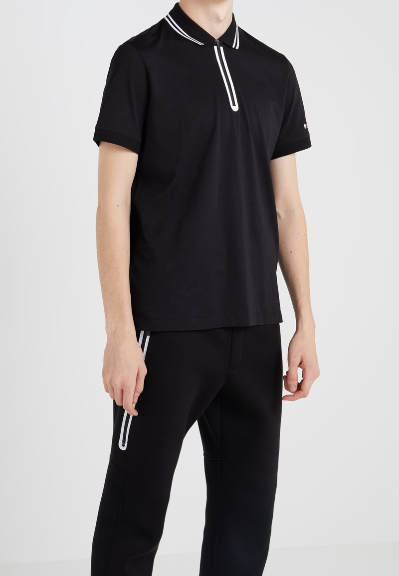 Homme portant un polo noir à manches courtes avec une bande blanche sur le col et une fermeture éclair, associé à un pantalon noir avec un détail de bande blanche.