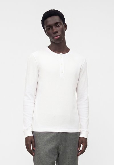 Jeune homme à la peau foncée portant un t-shirt henley blanc à manches longues et un pantalon gris, debout devant un fond clair uni.