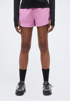 Nogi osoby ubranej w różowe sportowe szorty, czarną bluzkę z długim rękawem, czarne skarpety do kostek oraz szare buty trekkingowe stojącej na białym, jednolitym tle.