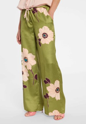 Femme portant un pantalon vert à jambes larges avec un grand imprimé floral crème et violet, assorti à des talons ouverts roses clairs et un haut rose.