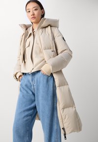 Lange beige pufferjas met een capuchon, voorzien van elastische accenten, zijzakken en een gladde glans, te dragen over een beige top en blauwe jeans.
