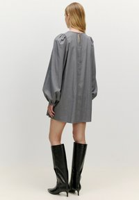 Graues kurzes Kleid mit Puffärmeln, das mit einem Schlüsselloch am Rücken und einem abgerundeten Saum versehen ist. Kombiniert mit schwarzen kniehohen Stiefeln mit Absatz.