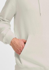 Lys grå hættetrøje med en frontlomme. En hånd ligger i lommen og viser en afslappet og uformel stil. Blød stofstruktur synlig.
