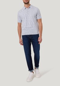 Man in een lichtgrijze polo met korte mouwen, donkerblauwe gekraagde jeans en witte sneakers, staand tegen een effen lichte achtergrond.