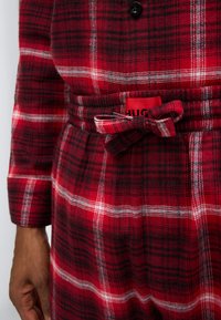 Κόκκινο πουκάμισο από καρό ύφασμα με κουμπιά, στενή μέση και ζώνη στο ίδιο σχέδιο, με μοτίβα σε μαύρο και λευκό και ορατή ετικέτα λογοτύπου.