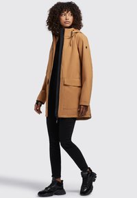 Giacca impermeabile marrone con cappuccio, zip frontale e due grandi tasche. Abbinata a pantaloni neri e scarpe da ginnastica nere. Tessuto liscio.