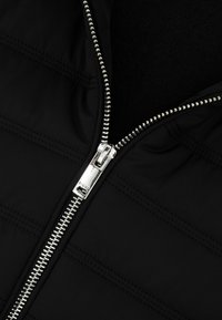 Srebrny metalowy zamek błyskawiczny częściowo zapięty na czarnej pikowanej tkaninie z widocznym przeszyciem.