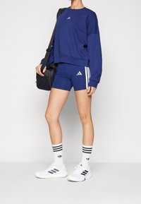 Felpa blu navy e pantaloni corti, con strisce e loghi bianchi. Scarpe da ginnastica bianche con dettagli neri e calze a strisce completano il look.