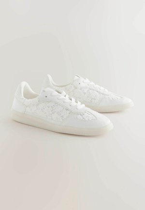 Valged spordijalatsid, millel on külgedel lipsupaneelid, sile nahakujuline materjal, ümar varvas ja tekstureeritud kummist tallad.