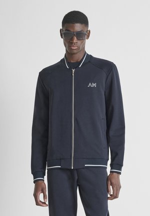 Hombre vestido con chaqueta de cremallera azul marino oscuro y pantalones a juego, con gafas de sol negras, de pie contra un fondo claro y liso.