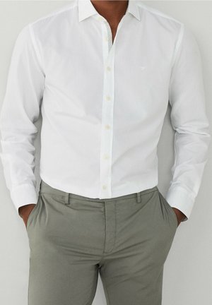 Homme portant une chemise blanche ajustée boutonnée, rentrée dans un pantalon gris clair, les mains dans les poches, sur un fond uni.