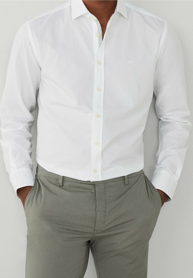 Homme portant une chemise blanche ajustée boutonnée, rentrée dans un pantalon gris clair, les mains dans les poches, sur un fond uni.