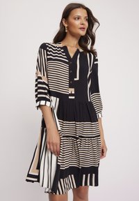 Czarna i kremowa sukienka w paski z dekoltem w serek zapinanym na guziki, z rękawami o długości trzech kwartałów i falbanowym dołem; posiada różnorodne wzory i tekstury pasków.