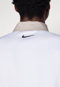 Camisola atlética branca com colarinho bege e logótipo preto da Nike nas costas. Textura suave, design minimalista, adequada para vestuário desportivo.