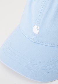Lyseblå caps laget av teksturert stoff, med en fremtredende hvit brodert logo. Buet skygge og synlige sømdetaljer.