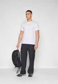 Camiseta blanca de manga corta con texto azul, pantalones cargo negros con bolsillos, mochila oscura y calzado gris/azul. Modelo de pie contra un fondo sencillo.