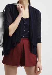 Marinefarbener transparenter Cardigan mit aufgerollten Ärmeln über einem marinefarbenen Oberteil mit kleinen Verzierungen. Dazu hochgeschnittene kastanienbraune Shorts. Silberne Tasche.