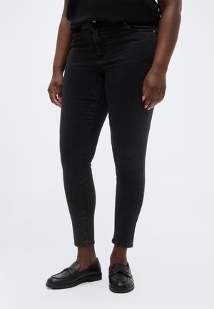 Άτομο που φοράει εφαρμοστά μαύρα τζιν και μαύρα δερμάτινα μοκασίνια, όρθιο μπροστά σε απλό ανοιχτό φόντο.